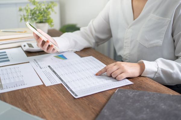 a woman at a desk looking at spreadsheets and comparing the data with content on her iphone.